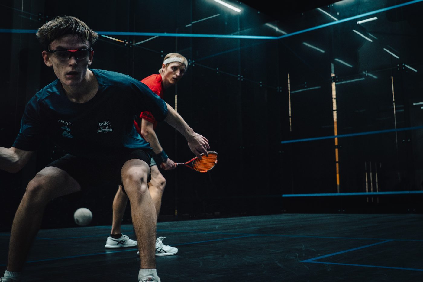 Two players in a hard rally on the glass court at YOLO Squash