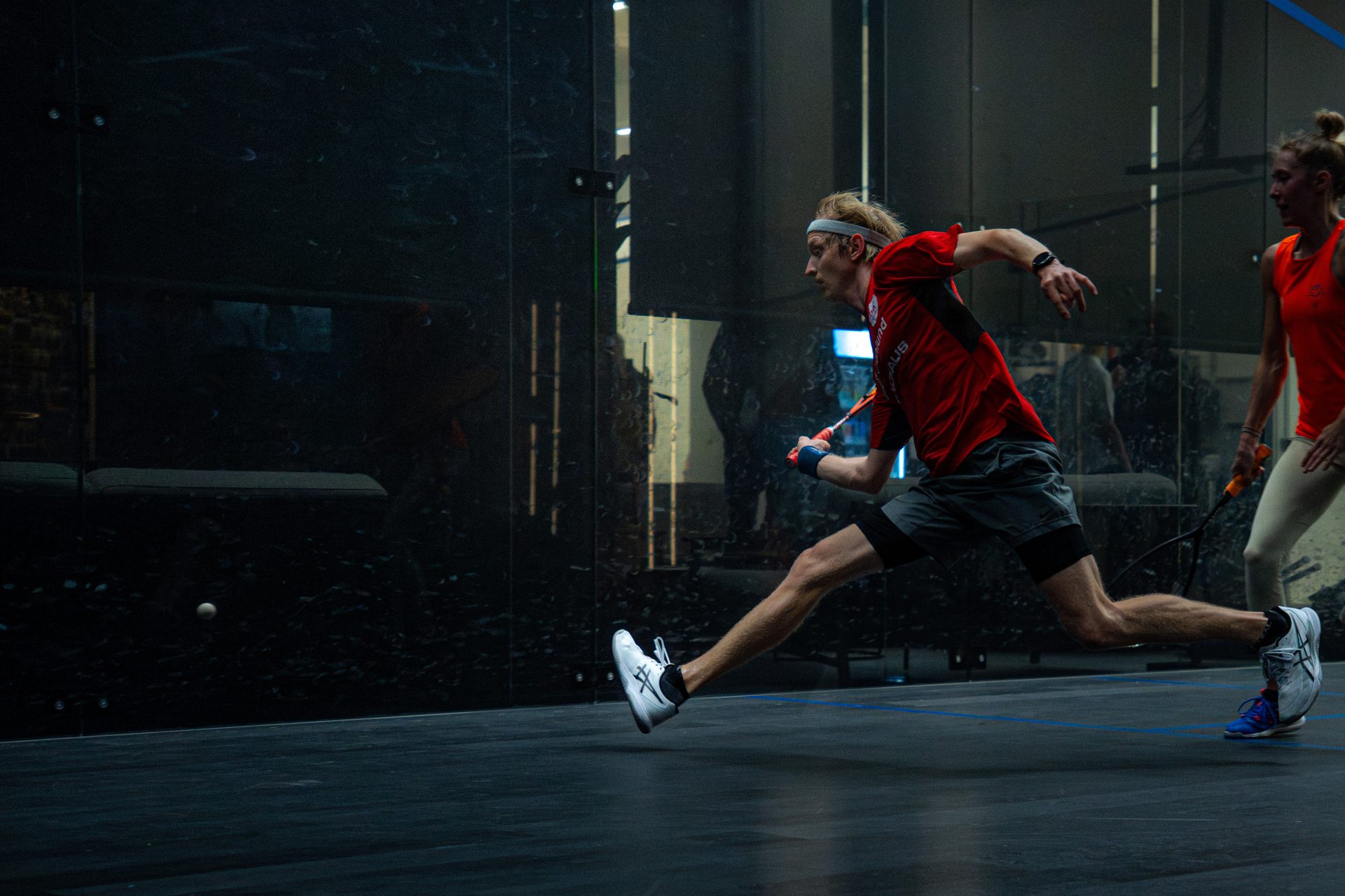 Squash player lunging to strike the ball on a glass court